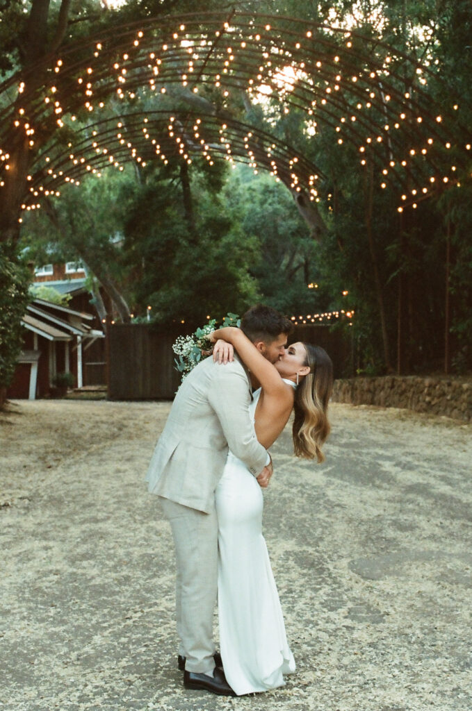 bride in satin halter style wedding dress and groom in khaki plaid suit walk under arch at Calamigos Ranch