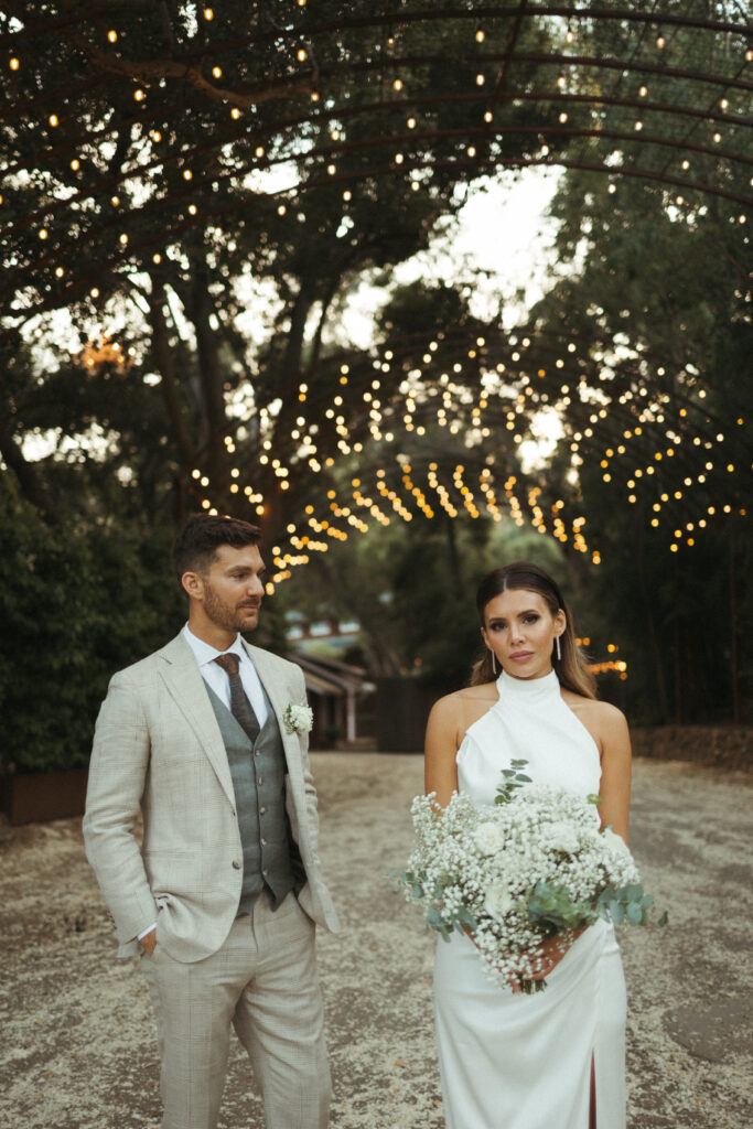 bride in satin halter style wedding dress and groom in khaki plaid suit walk under arch at Calamigos Ranch