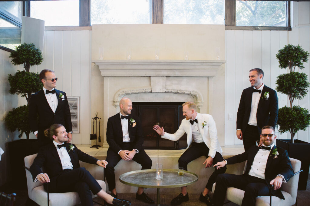 groom in white suit jacket and black bow tie sits on fireplace talking with wedding party in black suits
