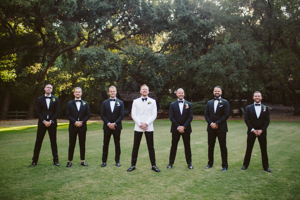 stylish groom in white tuxedo jacket and black bowtie stands with wedding party in black suits at Calamigos Ranch