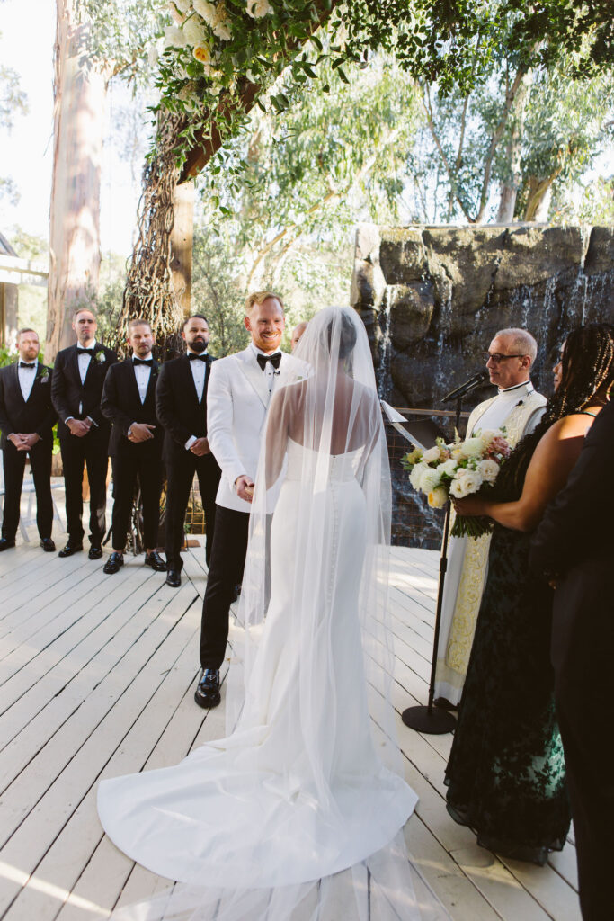 bride in cathedral length veil says vows to groom in white tuxedo jacket during wedding ceremony in front of a waterfall at Calamigos Ranch 