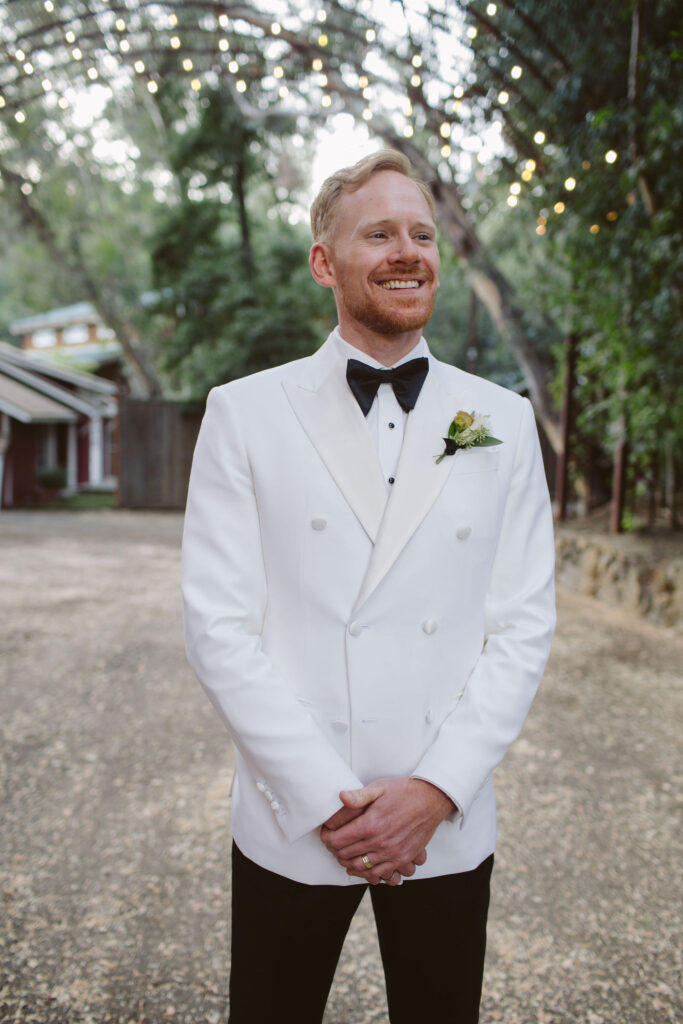 groom in white tuxedo jacket with black bowtie and pants and simple boutonniere 