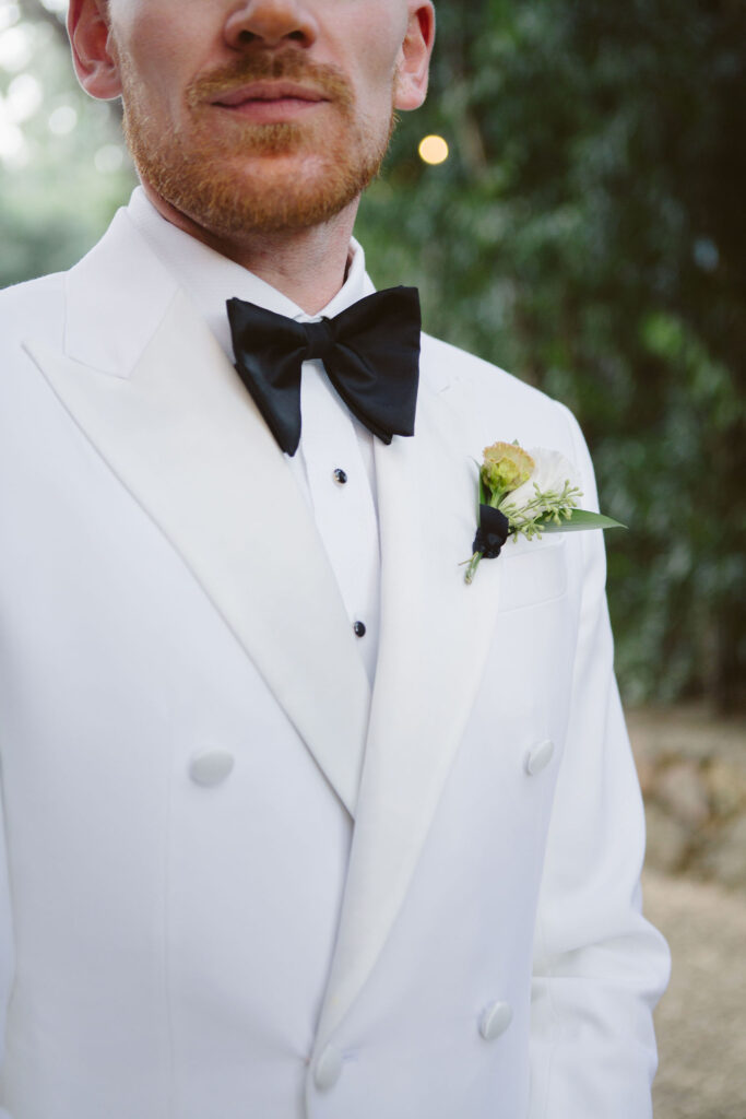 groom in white tuxedo jacket with black bowtie and pants and simple boutonniere 