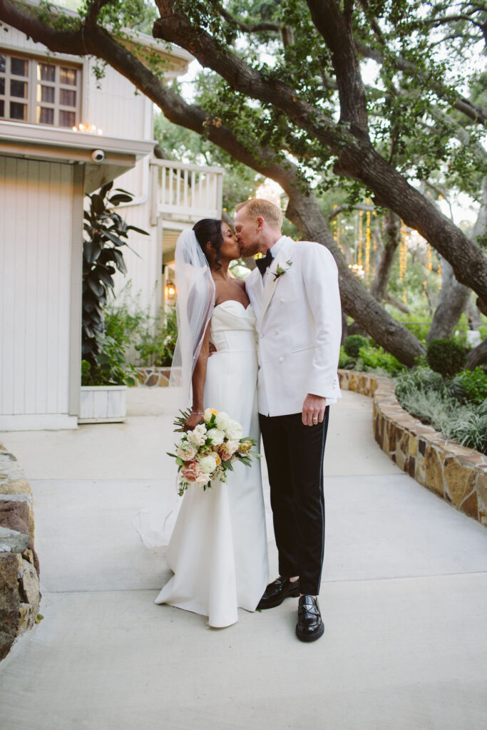 bride in modern minimalist wedding dress and cathedral veil kisses groom in white tuxedo jacket at Calamigos Ranch 
